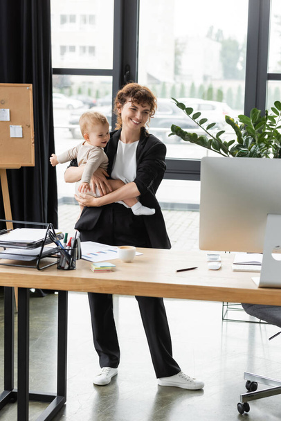 ευτυχής επιχειρηματίας με μαύρο κοστούμι κοιτάζοντας οθόνη υπολογιστή, ενώ στέκεται με μικρό παιδί στο γραφείο - Φωτογραφία, εικόνα