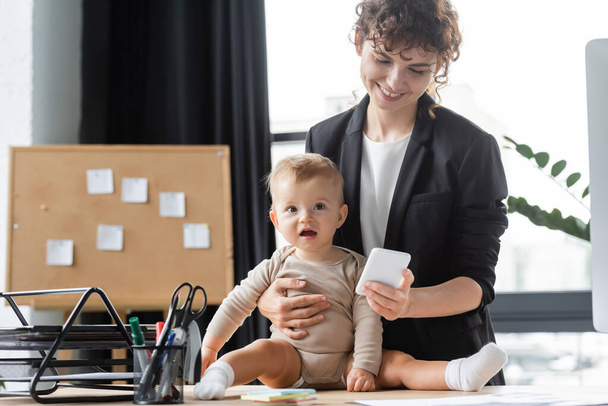 μικρό μωρό στο romper κάθεται στο γραφείο κοντά χαμογελαστή μαμά σε μαύρο σακάκι χρησιμοποιώντας το κινητό τηλέφωνο στο γραφείο - Φωτογραφία, εικόνα