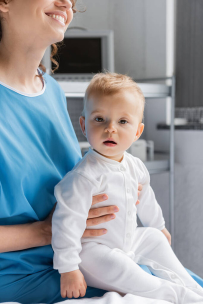 cheerful woman in patient gown holding little daughter looking at camera in clinic - Foto, immagini