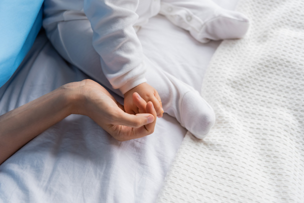 πάνω άποψη του περικοπή γυναίκα κρατώντας το χέρι του μικρού κοριτσιού σε romper κάθεται στο κρεβάτι του νοσοκομείου - Φωτογραφία, εικόνα