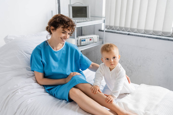 женщина в больничном платье улыбается рядом с маленькой дочерью на кровати в больничном отделении - Фото, изображение