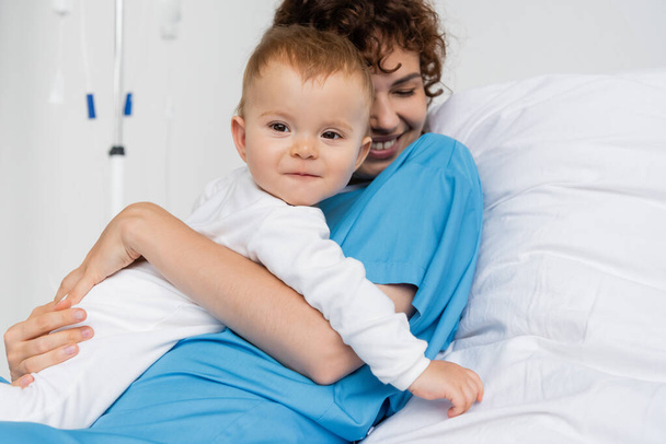 счастливая женщина в больничном платье обнимает маленькую дочь на кровати в клинике - Фото, изображение