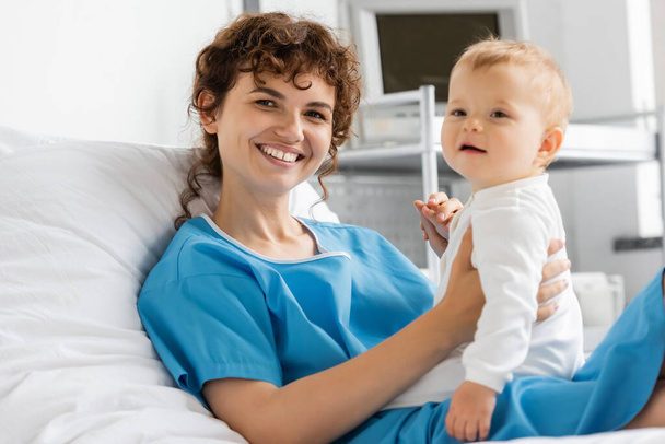 joyful woman in patient gown looking at camera near little daughter in hospital ward - Zdjęcie, obraz