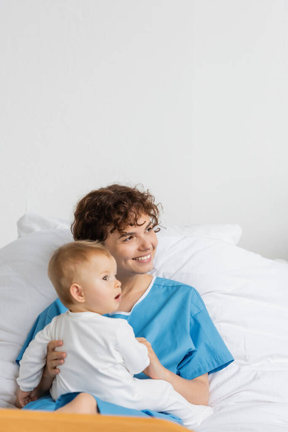 pleased woman and toddler girl with open mouth looking away on bed in hospital - Foto, Bild
