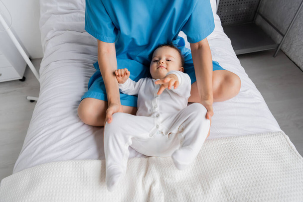 θέα υψηλής γωνίας της γυναίκας σε φόρεμα ασθενή κάθεται με σταυρωμένα τα πόδια κοντά στο μωρό σε romper στο κρεβάτι του νοσοκομείου - Φωτογραφία, εικόνα