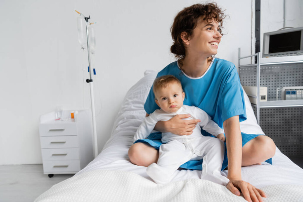 cheerful woman in patient gown looking away while sitting with toddler baby on hospital bed - Photo, image