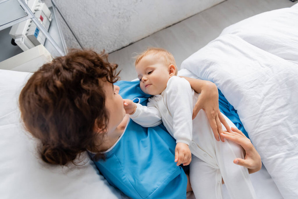 вид зверху на жінку в сукні пацієнта, що тримає дочку малюка на ліжку в лікарняному палаті
 - Фото, зображення