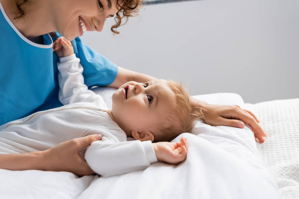 side view of happy woman looking at little girl in romper lying on bed in hospital - Fotó, kép