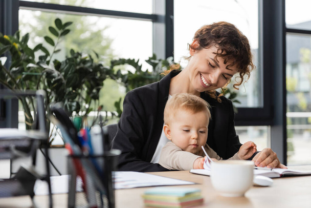 cheerful businesswoman sitting at workplace with toddler daughter near notebook and blurred coffee cup - Photo, image