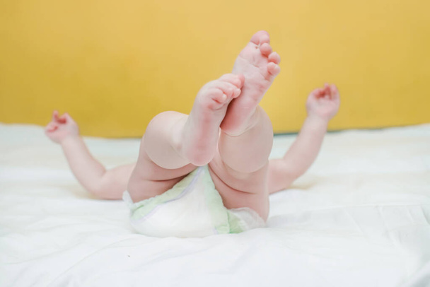 Vaaleanpunainen korkokengät pieni vauva makaa selällään valkoisella sängyllä ja leikkii omin käsin. Lapsesta huolehtiminen. Vaihda vaipat vastasyntyneille. - Valokuva, kuva