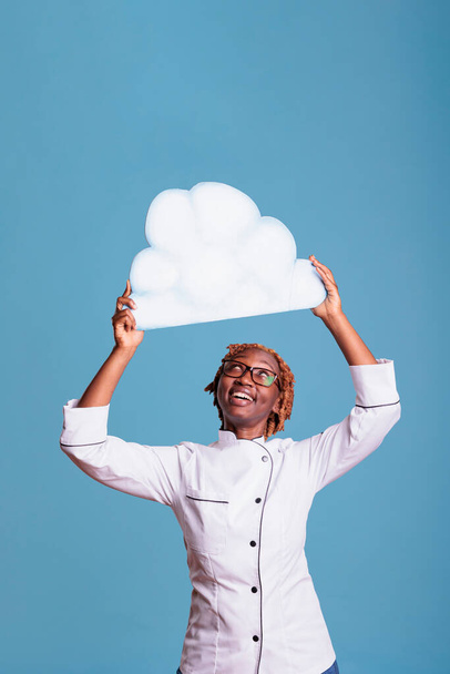 Cheerful woman wearing chef jacket holding cloud of text above head with space for advertising. Smiling cook with white text bubble in studio shot against blue background. - Foto, immagini