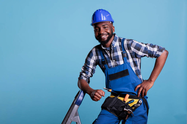 Builder looking happy and relaxed at work, dressed in coveralls and protective helmet leaning on portable ladder. Optimistic construction worker wearing tool belt while taking a break. - Foto, Imagen