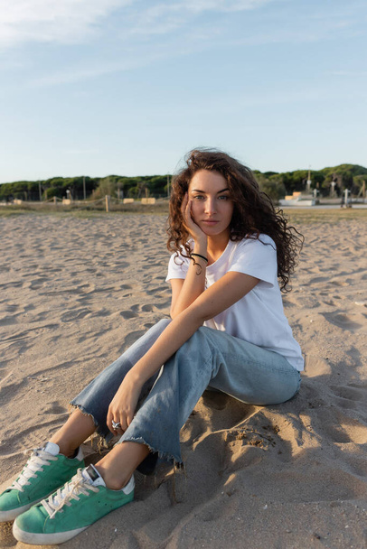 kudrnatá mladá žena sedící v džínách a bílém tričku na písečné pláži v Barceloně  - Fotografie, Obrázek