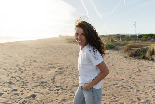 joyeux jeune femme en t-shirt blanc marchant sur la plage de sable en Espagne  - Photo, image