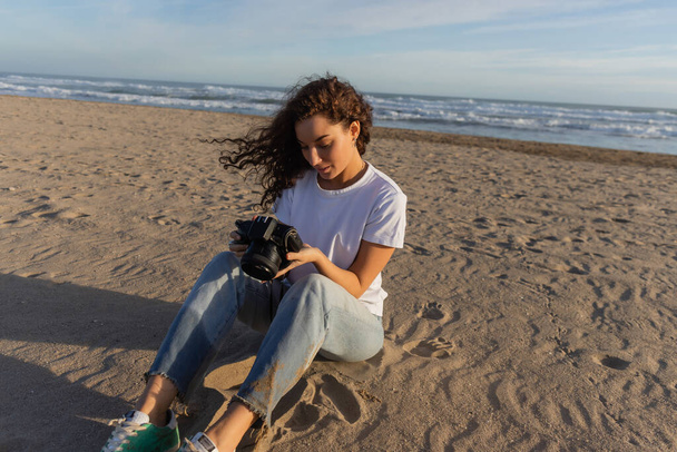 curly woman sitting in blue jeans and white t-shirt sitting with digital camera on beach in Spain  - Fotó, kép