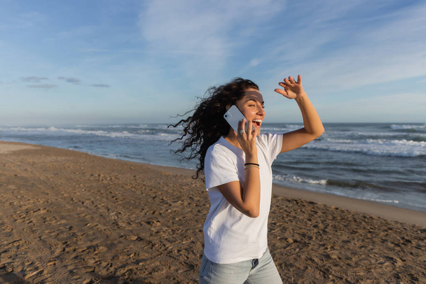 amazed woman in white t-shirt talking on smartphone on sandy beach in Barcelona  - Photo, image