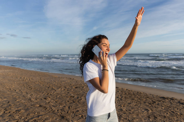 femme excitée en t-shirt blanc parlant sur smartphone et agitant la main sur la plage à Barcelone  - Photo, image