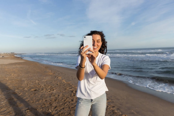 fröhliche Frau im weißen T-Shirt, die am Sandstrand von Barcelona ein Foto mit dem Smartphone macht  - Foto, Bild