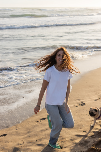 joyeux jeune femme courir avec chiot chiot sur la plage de sable fin pendant le coucher du soleil  - Photo, image