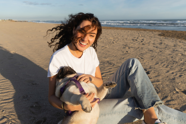 cheerful young woman with curly hair holding pug dog while sitting on sand near sea in Spain  - Valokuva, kuva