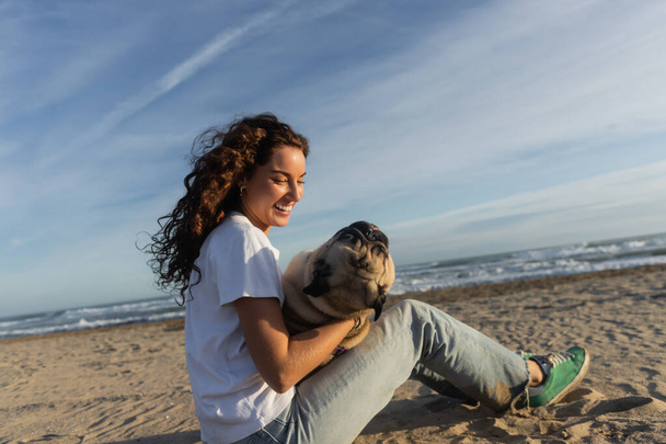 happy young woman with curly hair holding pug dog while sitting on beach near sea in Spain  - Fotografie, Obrázek