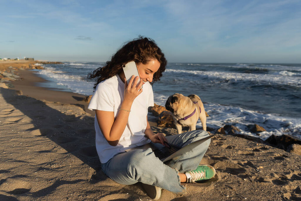 cheerful freelancer talking on smartphone while using laptop near pug dog on beach near sea in Spain  - Foto, Imagen