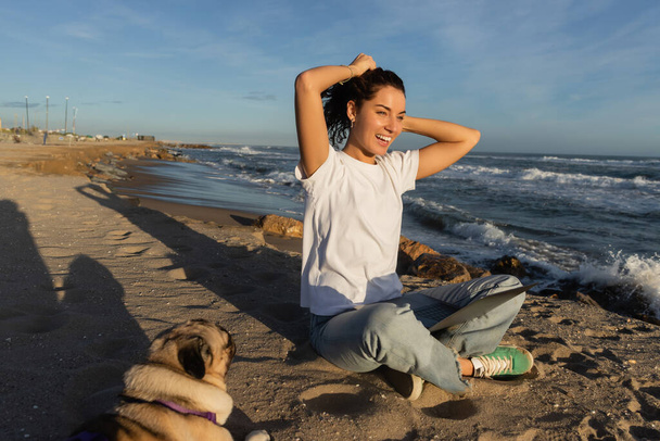 веселый фрилансер, сидящий с ноутбуком и регулирующий вьющиеся волосы рядом с собакой-мопсом на пляже возле моря в Испании  - Фото, изображение