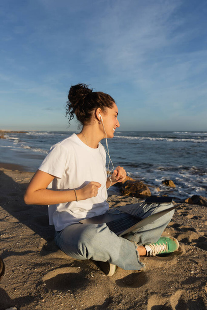 sonriente joven freelancer en auriculares con cable sentado con portátil en la playa de Barcelona - Foto, imagen
