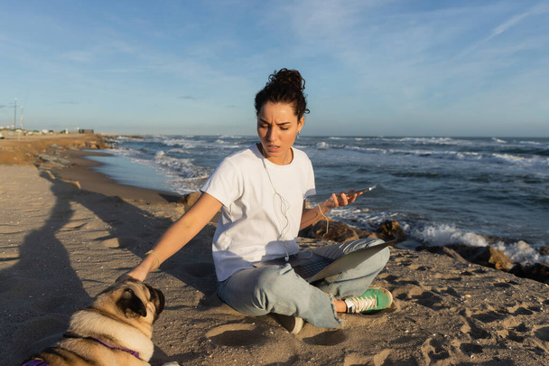 young woman in wired earphones holding smartphone near laptop and cuddling pug dog on beach in Barcelona - Photo, Image