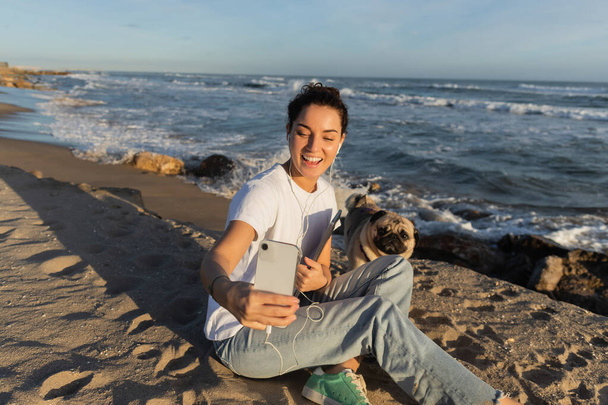 glückliche Frau mit kabelgebundenen Kopfhörern, Laptop in der Hand und Selfie mit Mops Hund am Strand von Barcelona - Foto, Bild