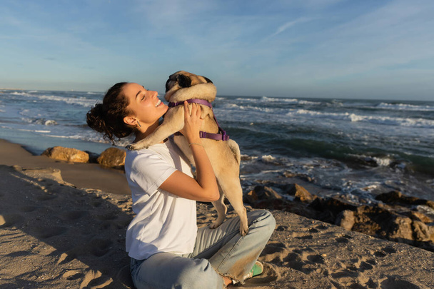 positive young woman with closed eyes holding pug dog on beach near sea in Barcelona  - Zdjęcie, obraz