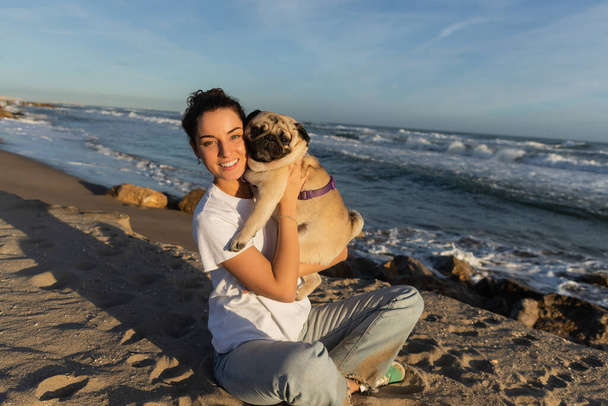 positieve jonge vrouw met krullend haar met pug hond op het strand in de buurt van zee in Barcelona  - Foto, afbeelding