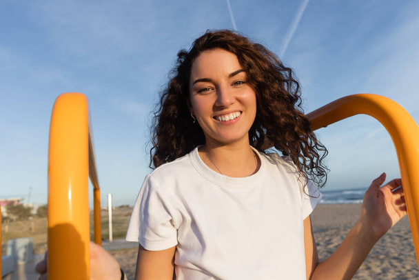 joyeuse jeune femme aux cheveux bouclés regardant la caméra près des mains courantes jaunes sur la plage de Barcelone  - Photo, image