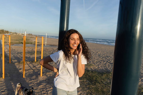 Barcelona sahilinde eli kalçasında dururken akıllı telefondan konuşan neşeli genç bir kadın.  - Fotoğraf, Görsel