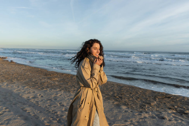 cheerful woman in stylish beige trench coat walking on beach in Barcelona  - Photo, Image