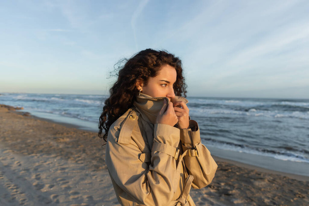curly young woman covering face with collar of beige trench coat on beach in Barcelona  - Foto, imagen
