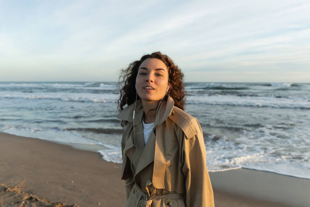 curly young woman in beige trench coat and wired earphones listening music near sea in Barcelona  - Foto, Bild