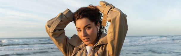 young woman in beige trench coat and wired earphones listening music while adjusting hair near sea in Barcelona, banner - Photo, image