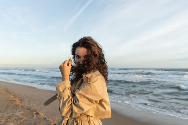 young woman in beige trench coat adjusting curly hair while looking at camera near sea in Barcelona  - Foto, Bild