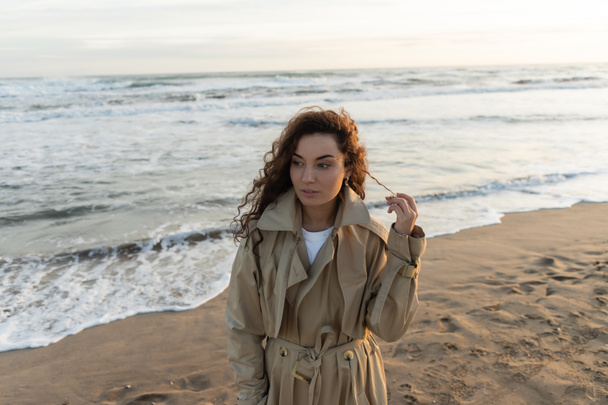 Young woman in trench coat touching curly hair on beach - Fotografie, Obrázek