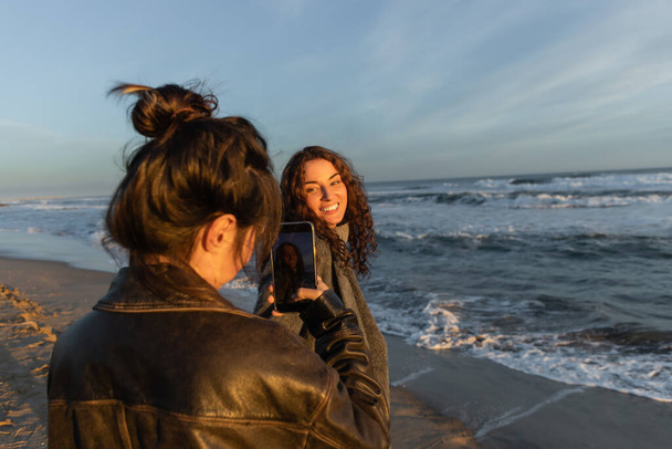 Woman taking photo on smartphone of smiling friend on beach in Barcelona  - Photo, Image