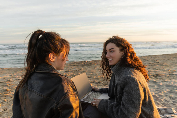 Улыбающаяся женщина с ноутбуком во время разговора с другом на пляже вечером  - Фото, изображение