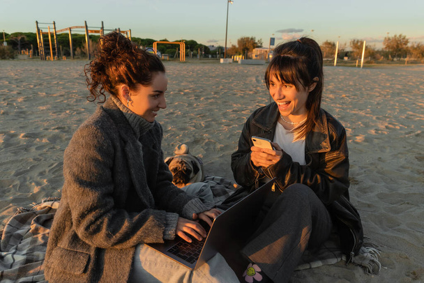 Cheerful women using gadgets near pug dog on beach in Spain  - Photo, Image