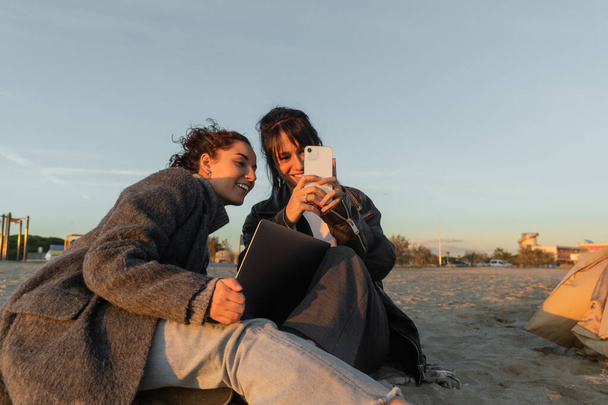 Smiling friends using smartphone and laptop on beach in Spain  - 写真・画像