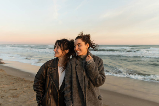 Mujeres jóvenes sonrientes mirando hacia otro lado mientras están de pie en la playa en España  - Foto, imagen