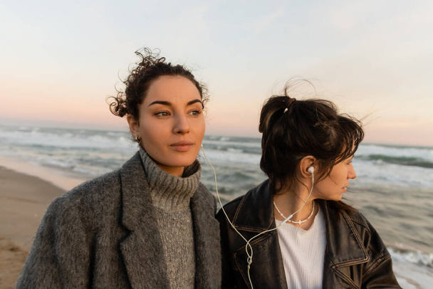 Curly woman listening music in wired earphones with friend on beach in Barcelona  - Фото, изображение