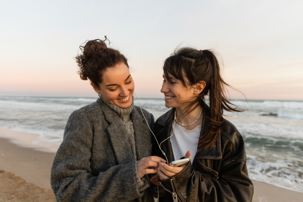 Hymyilevät ystävät kuuntelevat musiikkia langallisissa kuulokkeissa ja pitävät älypuhelinta rannalla   - Valokuva, kuva