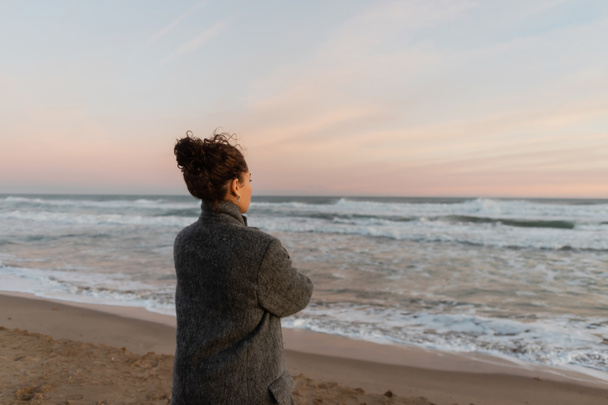 Göndör nő kabátban nézi a tengert, miközben áll a strandon Barcelonában  - Fotó, kép