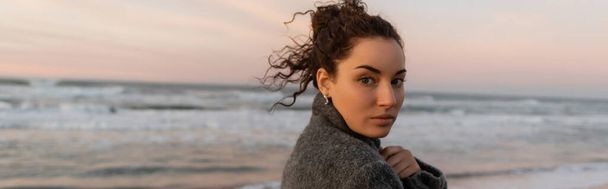Curly young woman in coat looking at camera with sea at background, banner  - Photo, Image