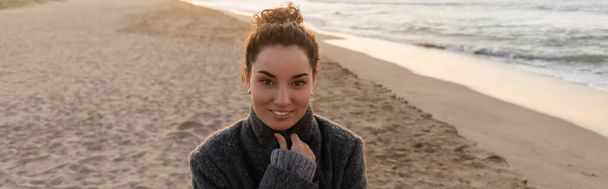 Smiling curly woman in coat looking at camera on beach in evening, banner  - Foto, immagini